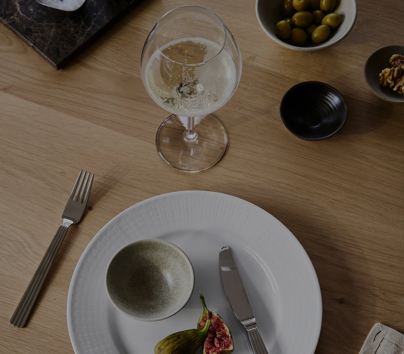 Um copo de champanhe, prato branco com uma tigela e figos, garfo, faca, azeitonas e nozes numa mesa de madeira.